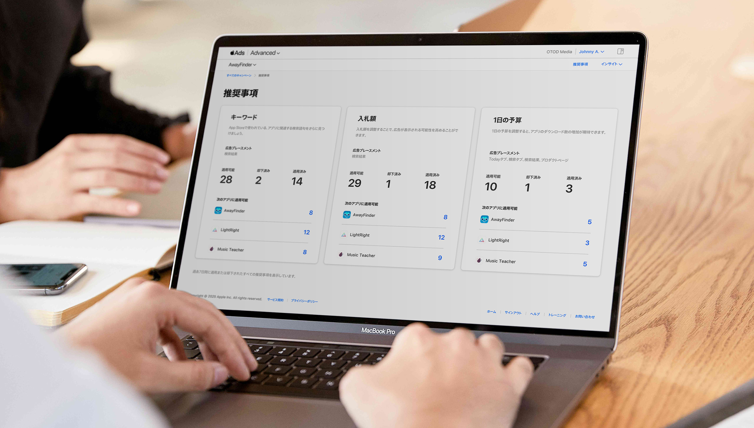 MacBookのキーボードで入力を行っている両手のクローズアップ。画面にはApple Adsの推奨事項ページが示され、キーワード、入札額、1日の予算ごとに整理された推奨事項が表示されている。