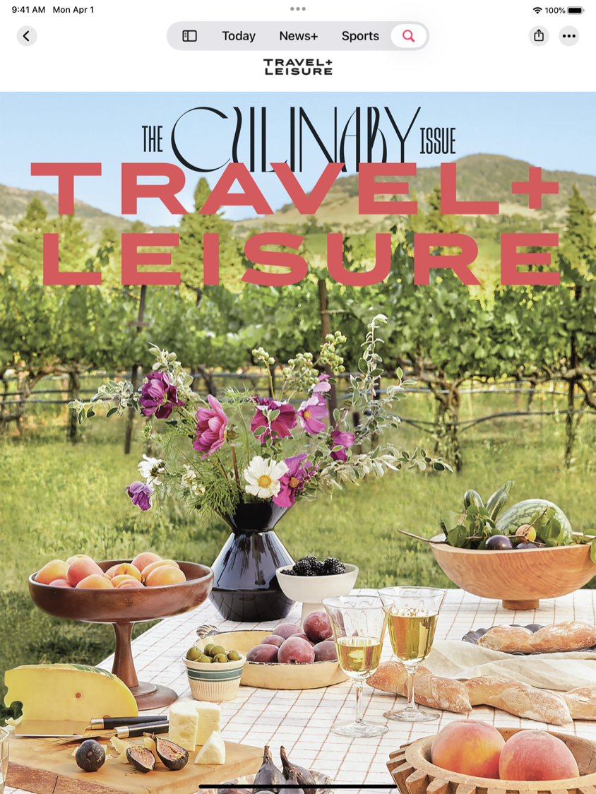 Das Cover von Travel + Leisure mit einem Bild eines mit Wein und Käse gedeckten Tisches mit einem Weinberg im Hintergrund.