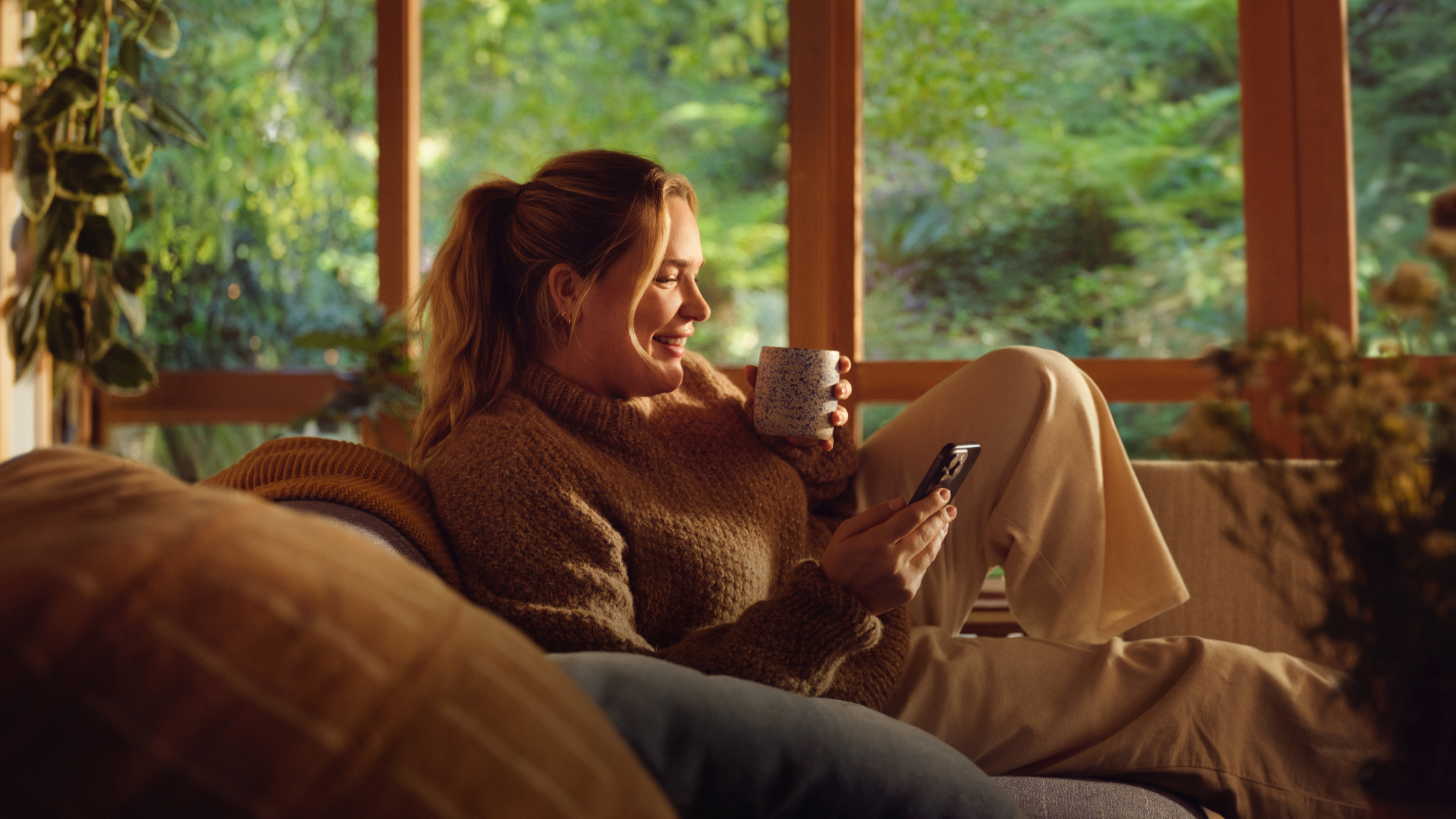 A woman sitting on her couch in the morning with a cup of coffee, smiling as she looks at her iPhone.
