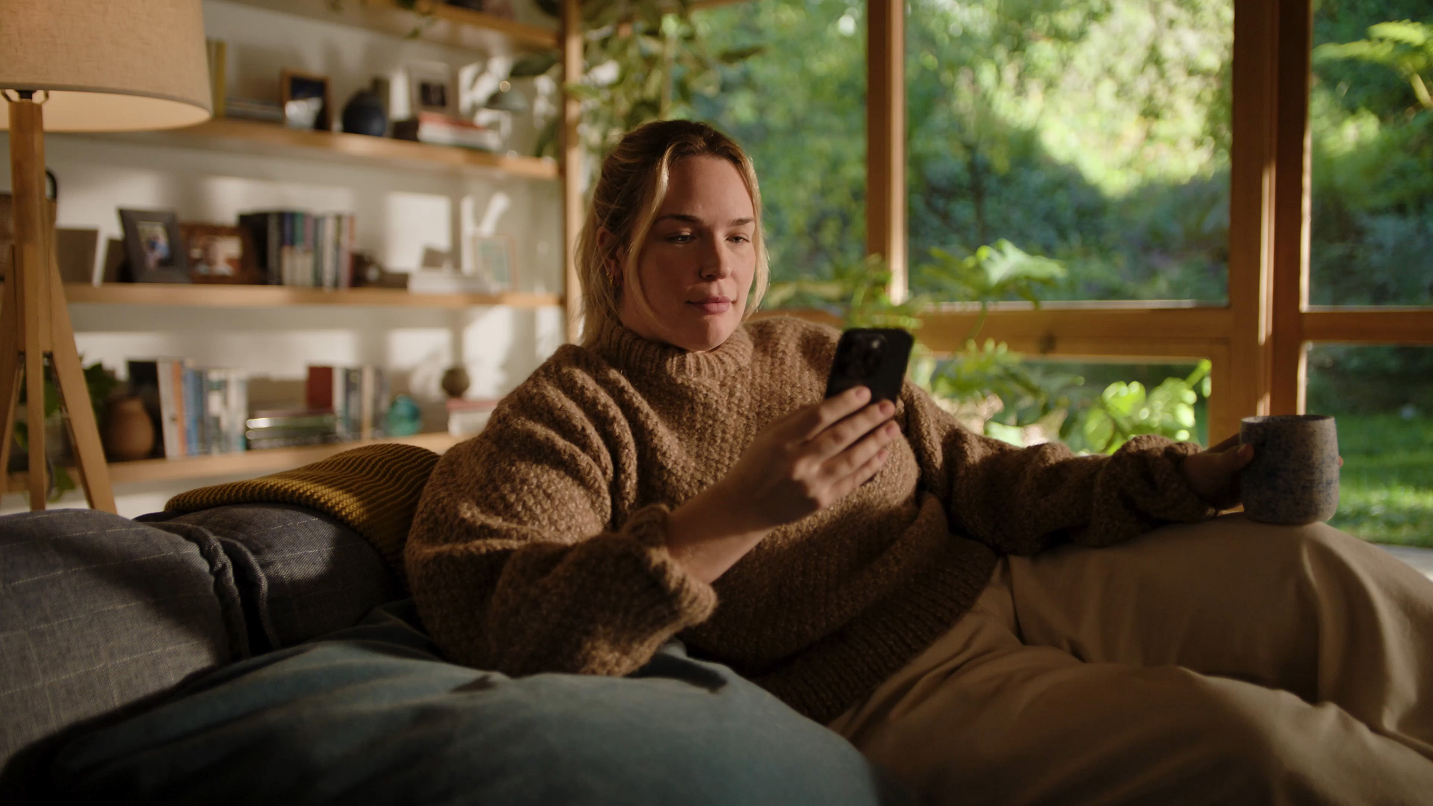  A woman sitting on her couch in the morning with a cup of coffee, smiling as she looks at her iPhone.